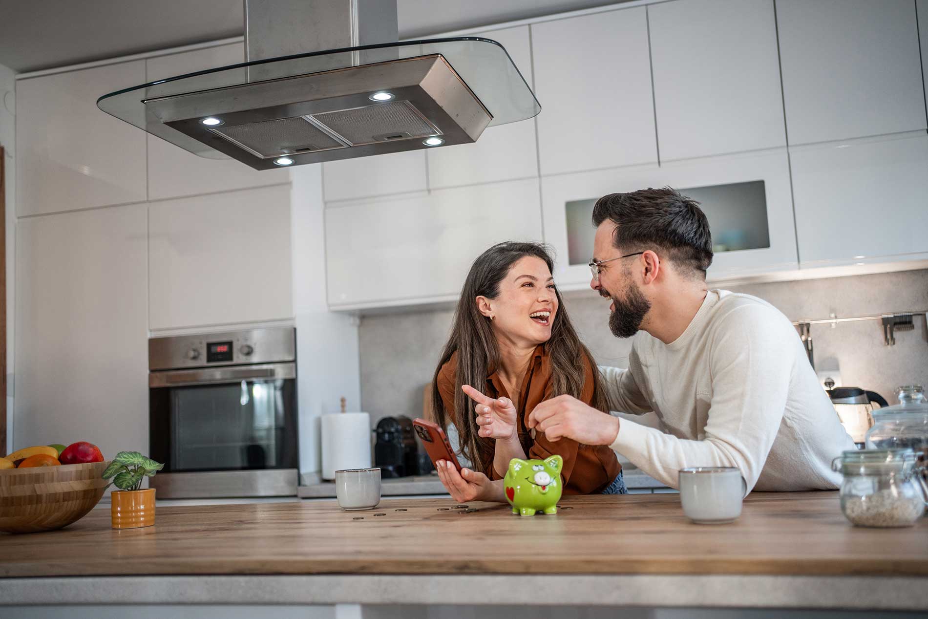 Couple laughing together at their kitchen bench beside a piggy bank