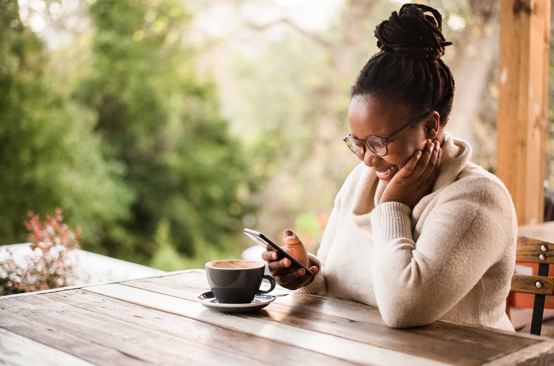Woman smiling as she reads a message on her phone over coffee on a verandah