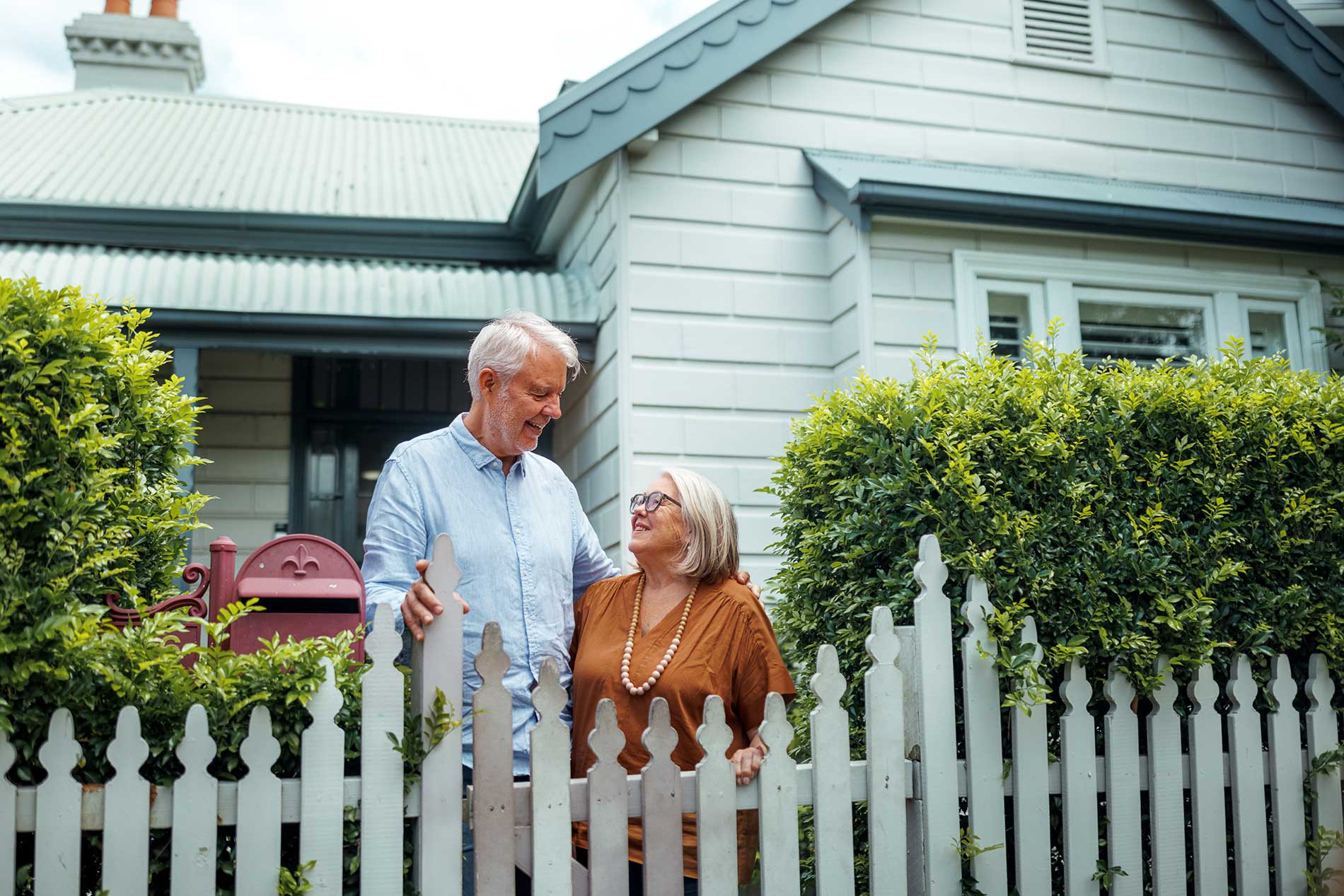 Retired couple smiling at each other at the front gate of their weatherboard home in Brisbane